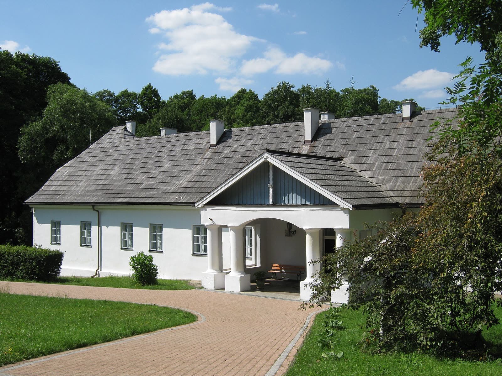 Biały parterowy dworek z gontowym dachem i kolumnowym gankiem, do którego prowadzi brukowana alejka, w letnim słońcu.