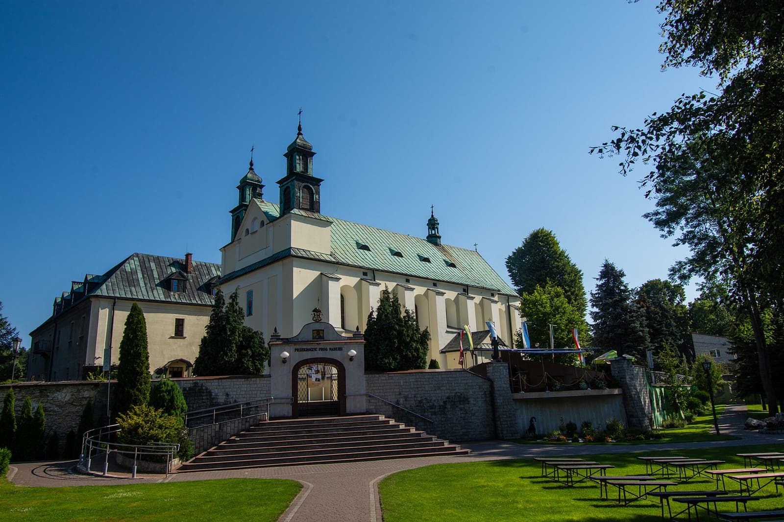 Biały kościół z dwiema wieżyczkami i klasztor paulinów w Leśniowie, a przed nimi schody prowadzące do bramy, w słoneczny dzień.