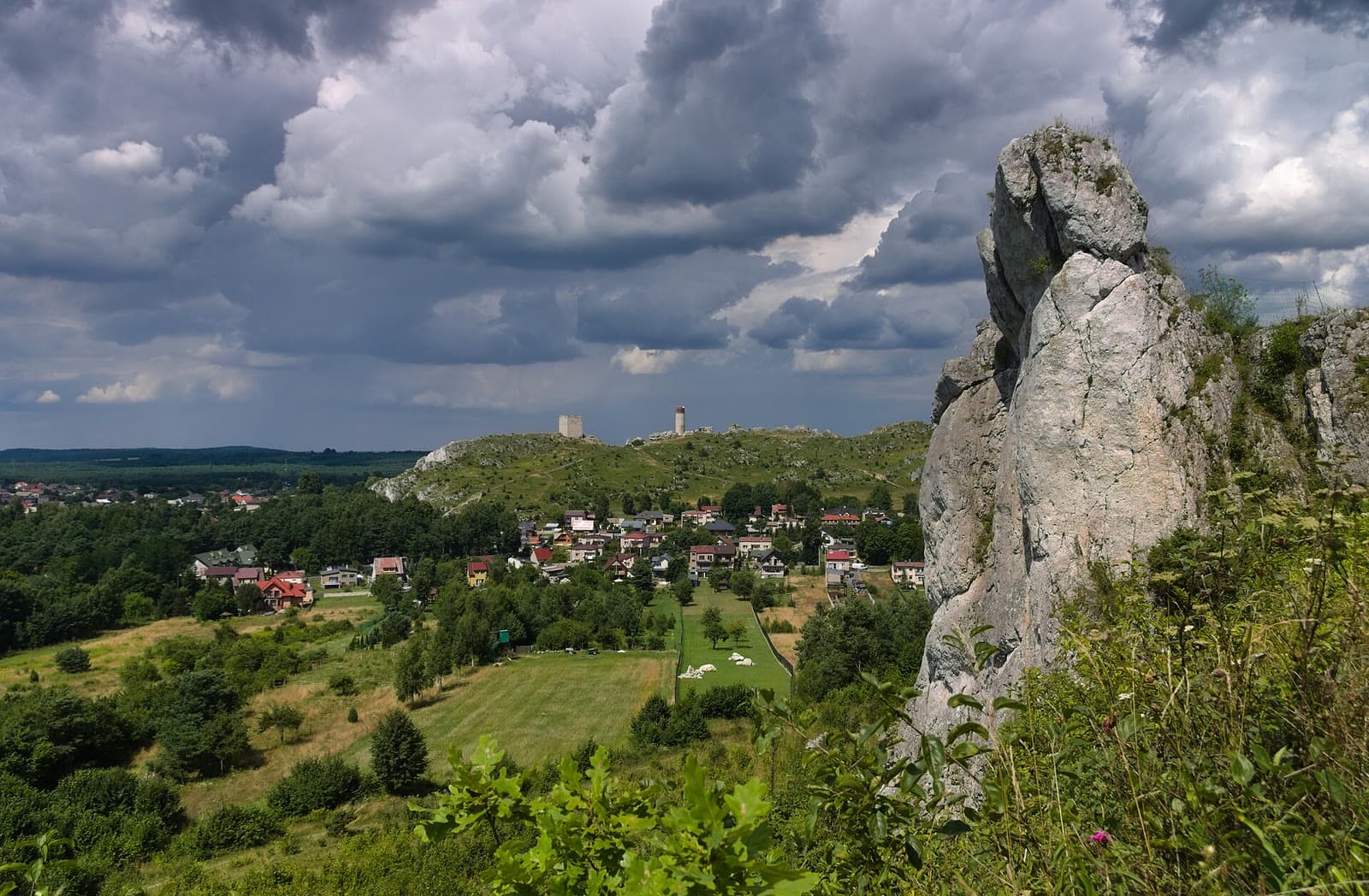Widok z Lipówek na Olsztyn i ruiny zamku na wzgórzu, z wapienną skałą Urwista Turnia po prawej i burzowymi chmurami nad krajobrazem Jury.