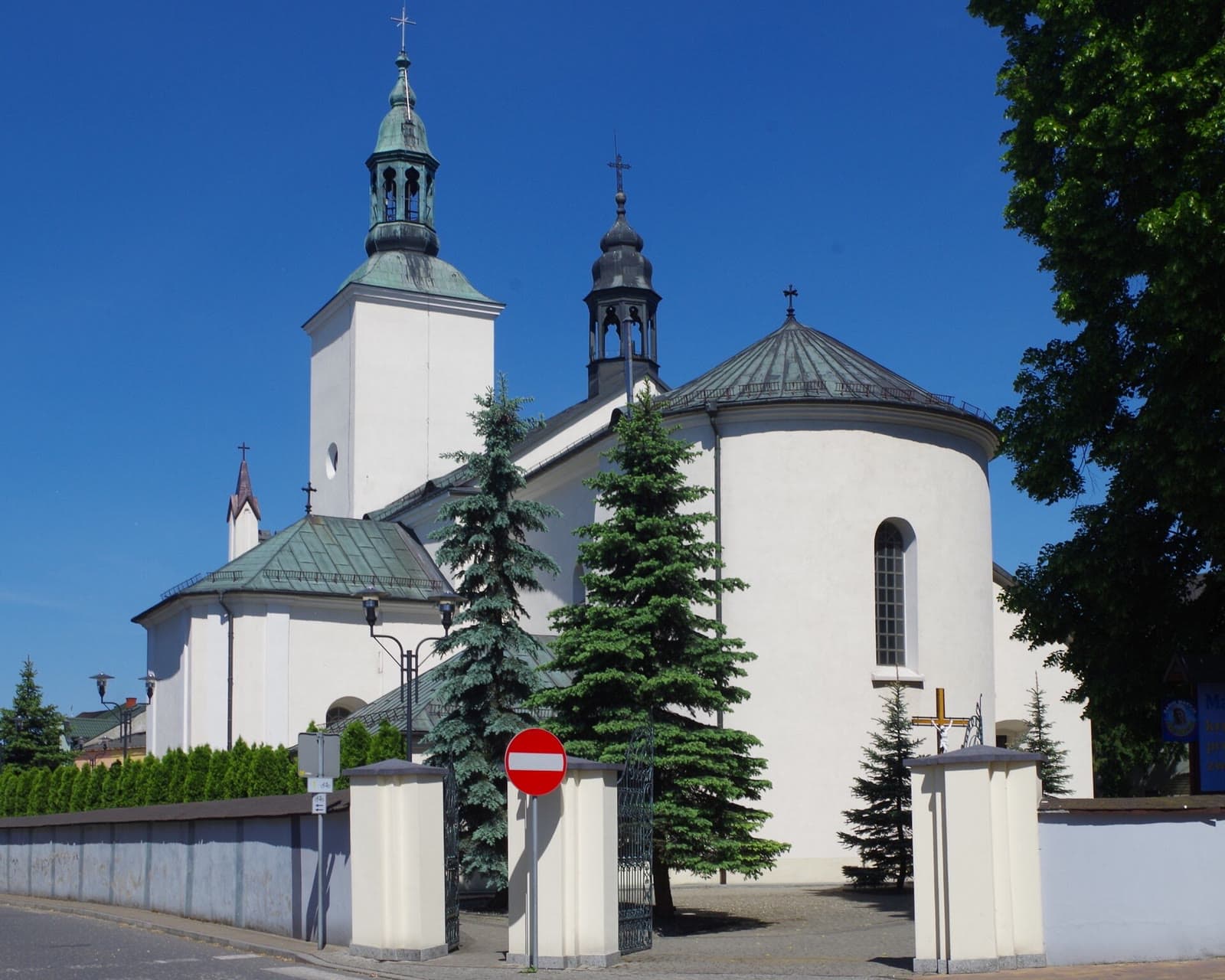 Biały kościół św. Apostołów Szymona i Judy Tadeusza w Żarkach z dwiema wieżami, otoczony zielenią, na tle błękitnego nieba.