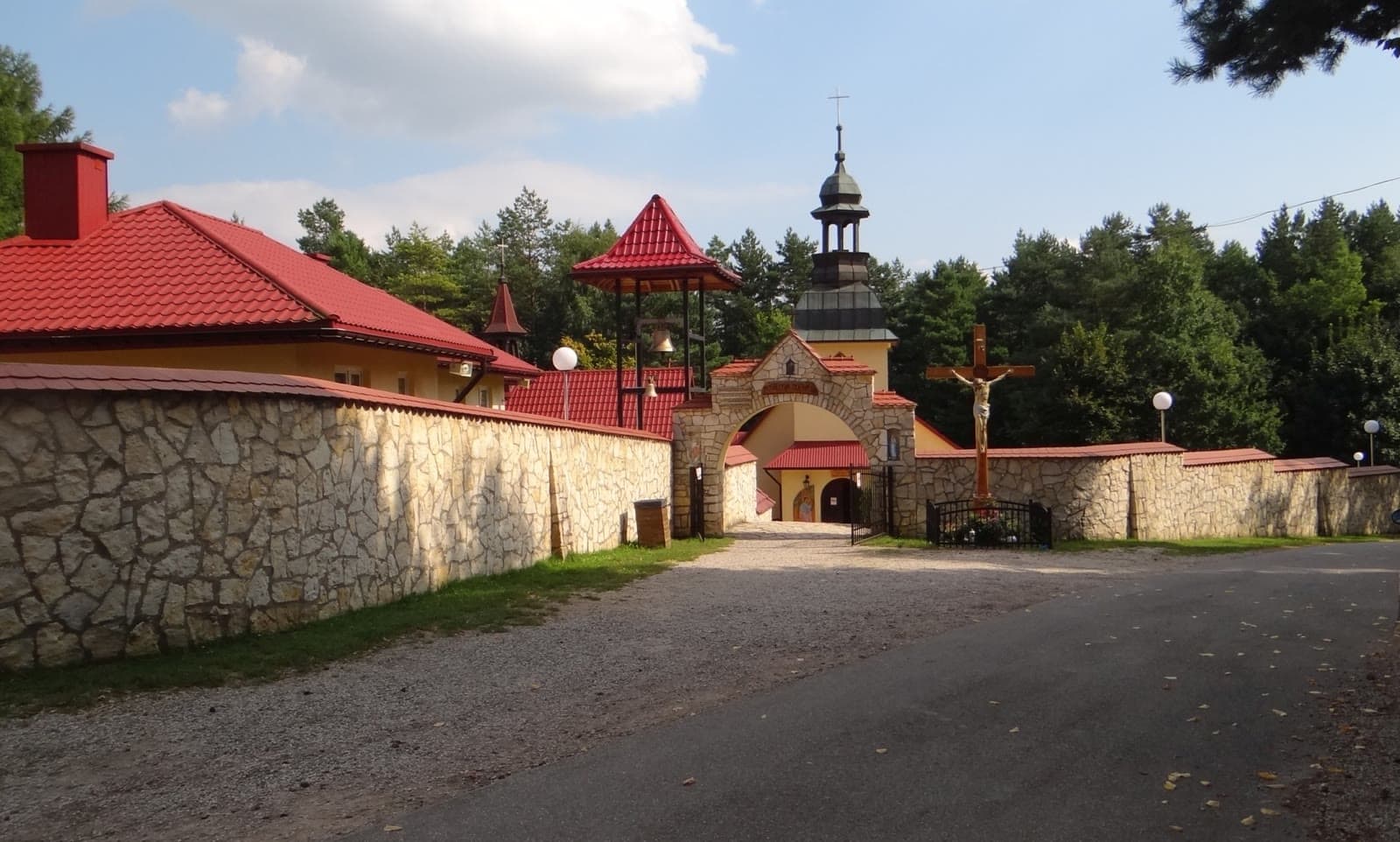 Kamienny mur z bramą, krzyż przydrożny i wieża kościoła dawnej pustelni w Czatachowej na tle sosnowego lasu.
